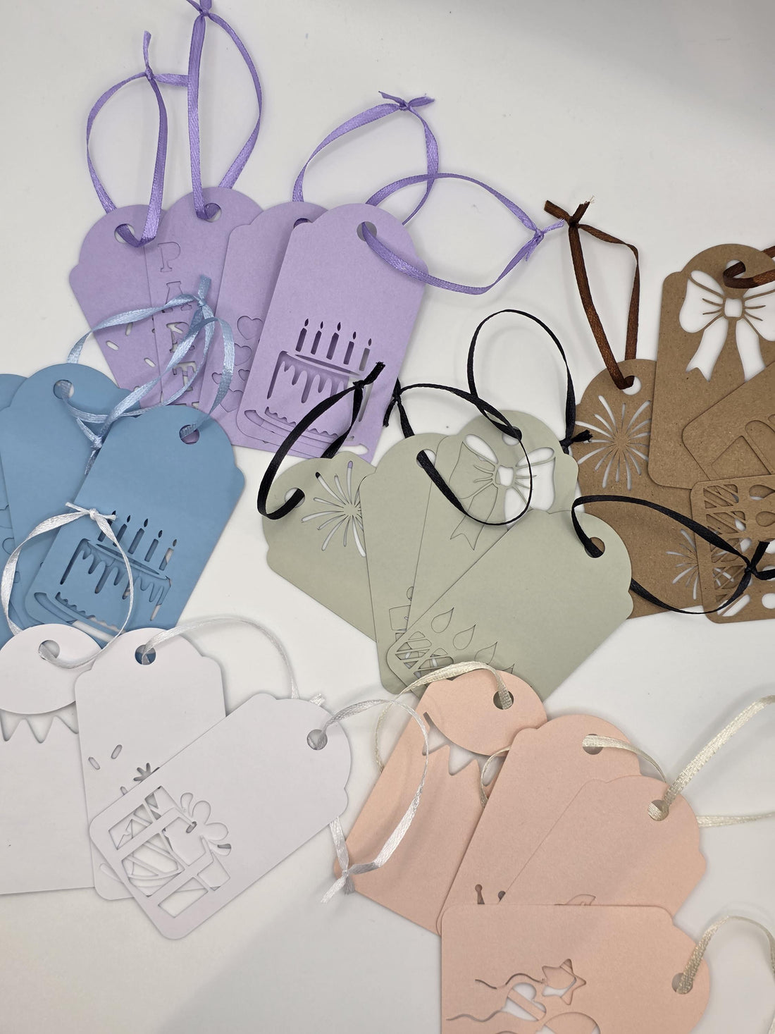 Sorted piles of die-cut cardstock gift tags in pink, white, blue, and sage green with attached satin ribbons.