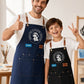 A full-length promotional photo of a smiling man and boy standing side-by-side in a brightly lit art studio with easels and paintbrushes visible in the background. Both wear white T-shirts, blue jeans, and matching heavy-duty canvas aprons with leather neck accents and large front pockets. The man on the left gives a thumbs up, wearing a dark blue apron featuring a circular white logo that reads "Breathe & Bloom Design" and a blue patch labeled "EVAN." The boy on the right poses with a peace sign, wearing a