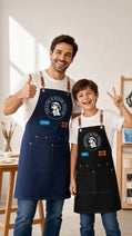 A full-length promotional photo of a smiling man and boy standing side-by-side in a brightly lit art studio with easels and paintbrushes visible in the background. Both wear white T-shirts, blue jeans, and matching heavy-duty canvas aprons with leather neck accents and large front pockets. The man on the left gives a thumbs up, wearing a dark blue apron featuring a circular white logo that reads "Breathe & Bloom Design" and a blue patch labeled "EVAN." The boy on the right poses with a peace sign, wearing a