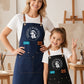 A full-length promotional photo of a smiling woman and young girl standing side-by-side in a sunlit art studio with wooden easels and paintbrushes in the background. Both are dressed in white T-shirts, blue jeans, and matching multi-pocket canvas aprons with leather strap hardware. The woman on the left has her hand on her hip, wearing a dark navy blue apron with the circular white "Breathe & Bloom Design" logo and a blue name patch reading "ELLIE." The girl on the right smiles while making a peace sign, we