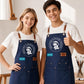 A full-length promotional photo of a smiling young woman and teenage boy standing side-by-side in a naturally lit art studio with wooden easels, canvases, and paintbrushes in the background. Both wear white T-shirts and matching navy blue canvas aprons equipped with brown leather neck straps, metallic grommets, and multiple utility front pockets. The woman on the left rests her hand on her hip, wearing an apron customized with the circular white "Breathe & Bloom Design" crest and a cyan patch reading "ELLIE