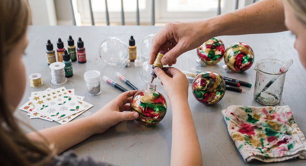 A close-up lifestyle shot of a person's hands and a child's hands decorating a clear glass ornament with colourful glitter and metallic inks. The scene is set on a grey wooden table surrounded by craft supplies, including small bottles of paint, glitter jars, markers, and other finished ornaments.