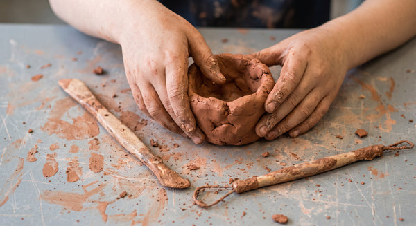 A close-up of a child's hands shaping a small clay bowl on a grey workbench with clay tools and clay residue.