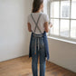 A full-length back view of a woman standing on hardwood floors in a bright, minimalist studio space facing a large multi-pane window. She wears a white crew-neck T-shirt, blue denim jeans, and white sneakers, holding a mug in one hand. She is wearing a blue apron highlighting its ergonomic cross-back design, featuring wide light-gray cotton webbing straps that crisscross over the back and thread through metallic grommets to tie in a neat bow at the waist.  