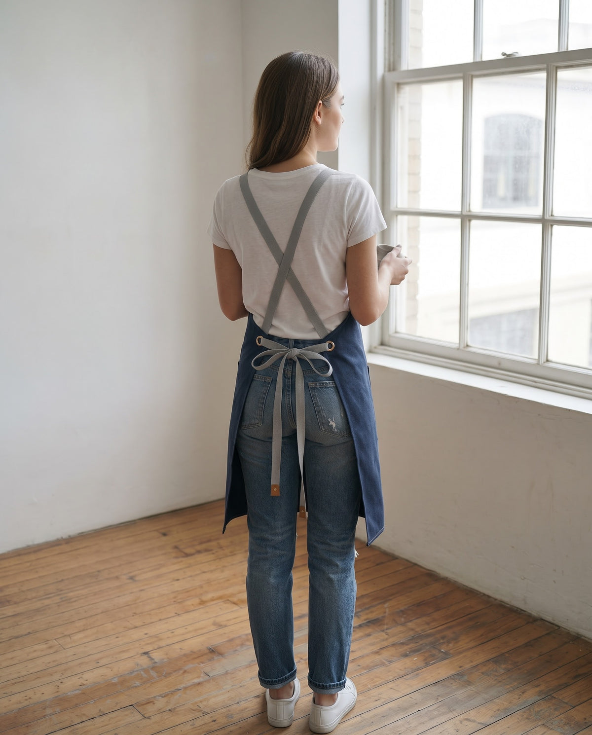 A full-length back view of a woman standing on hardwood floors in a bright, minimalist studio space facing a large multi-pane window. She wears a white crew-neck T-shirt, blue denim jeans, and white sneakers, holding a mug in one hand. She is wearing a blue apron highlighting its ergonomic cross-back design, featuring wide light-gray cotton webbing straps that crisscross over the back and thread through metallic grommets to tie in a neat bow at the waist.  
