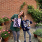 A smiling woman and young girl standing in a brick courtyard garden, holding up their textured abstract mixed-media canvas paintings.