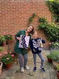 A smiling woman and young girl standing in a brick courtyard garden, holding up their textured abstract mixed-media canvas paintings.