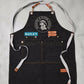 A top-down, flat lay product shot of a black heavy-duty canvas work apron laid flat against a neutral concrete background. The apron features brass grommets at the top bib and waist, fitted with detachable brown leather hardware and long light-gray cotton webbing straps. Centered on the chest is a round white printed logo reading "Breathe & Bloom Design," positioned directly above a cyan blue rectangular patch personalized with the name "BAILEY" in white capital letters. Double-stitched utility compartments