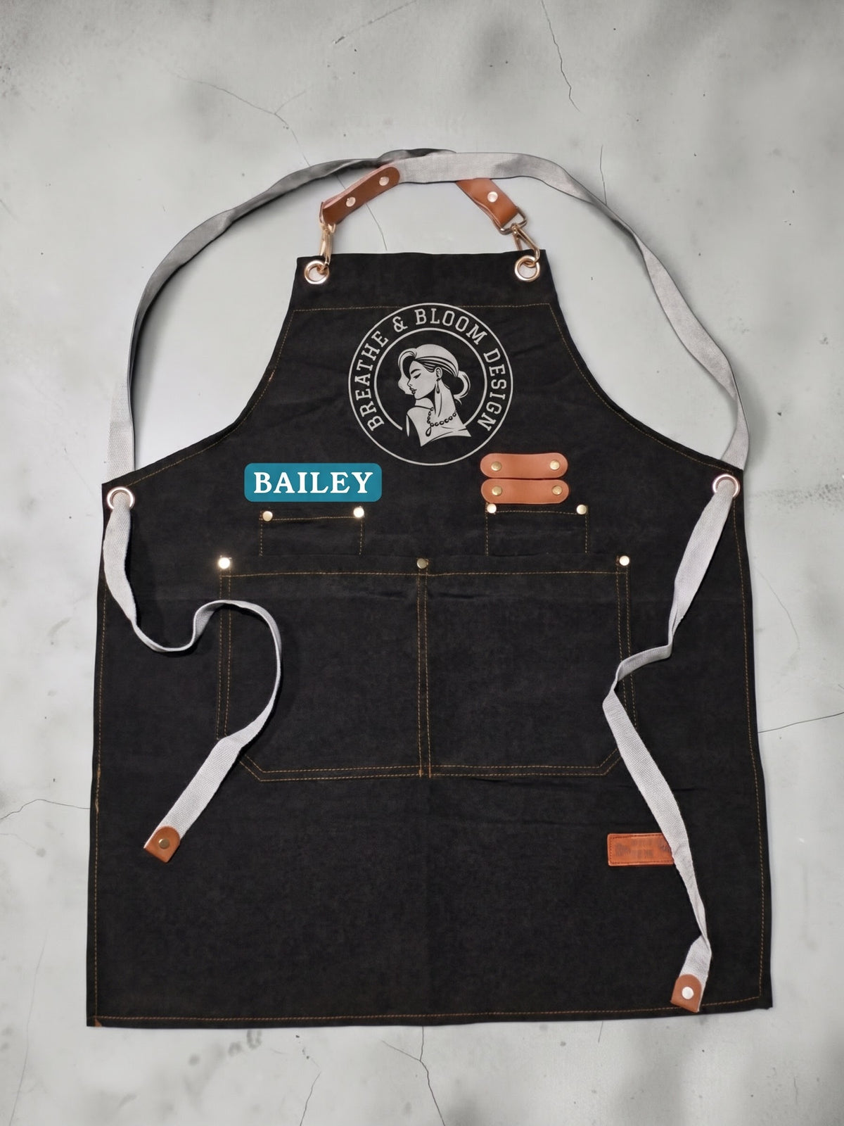A top-down, flat lay product shot of a black heavy-duty canvas work apron laid flat against a neutral concrete background. The apron features brass grommets at the top bib and waist, fitted with detachable brown leather hardware and long light-gray cotton webbing straps. Centered on the chest is a round white printed logo reading "Breathe & Bloom Design," positioned directly above a cyan blue rectangular patch personalized with the name "BAILEY" in white capital letters. Double-stitched utility compartments