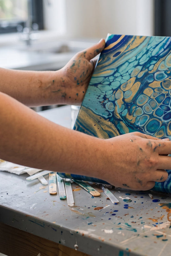 A person in a paint-splattered apron holds a square canvas featuring a fluid pour painting with an abstract pattern in blue, gold, and white. The workspace includes paint tubes, brushes in a glass jar, and splattered surfaces around the table.