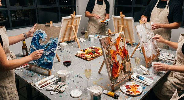 A group of people wearing beige aprons paint on canvases set up on easels around a table filled with art supplies, wine glasses, and a platter of fruit and cheese in a studio.