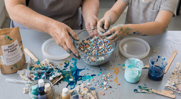 A person and a child mixing colorful decorative craft pieces in a glass bowl on a table covered with paint splatters, alongside a bag labeled white eco-friendly powder and small paint containers.
