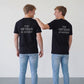  Two young men viewed from behind, standing side-by-side in a brightly lit studio. They wear matching black crewneck t-shirts featuring the stacked white text "THE ARTISAN ACADEMY" on the upper back, paired with light-wash blue jeans and white canvas sneakers. The man on the right, with short blonde-brown hair, candidly laughs while placing a friendly arm on the other’s shoulder.