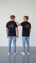  Two young men viewed from behind, standing side-by-side in a brightly lit studio. They wear matching black crewneck t-shirts featuring the stacked white text "THE ARTISAN ACADEMY" on the upper back, paired with light-wash blue jeans and white canvas sneakers. The man on the right, with short blonde-brown hair, candidly laughs while placing a friendly arm on the other’s shoulder.