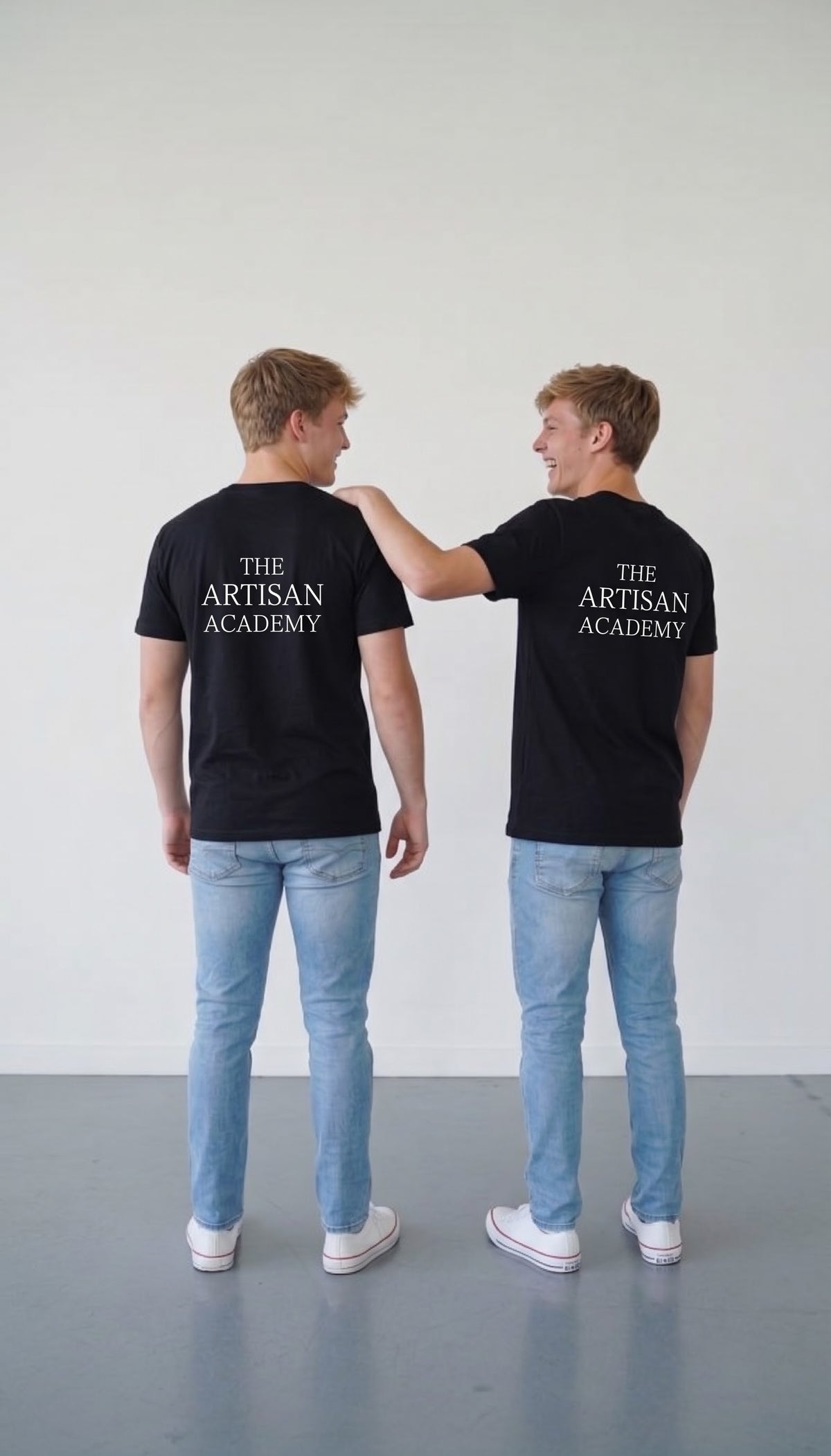  Two young men viewed from behind, standing side-by-side in a brightly lit studio. They wear matching black crewneck t-shirts featuring the stacked white text "THE ARTISAN ACADEMY" on the upper back, paired with light-wash blue jeans and white canvas sneakers. The man on the right, with short blonde-brown hair, candidly laughs while placing a friendly arm on the other’s shoulder.