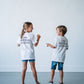 A young girl and a boy viewed from behind, captured in a candid, playful moment indoors. Both wear matching white crewneck t-shirts featuring the stacked dark text "THE ARTISAN ACADEMY" on the upper back. The girl on the left, with her hair in a ponytail, wearing denim shorts and patterned pink sneakers, gestures towards the boy on the right, who wears longer denim shorts and dark sneakers as they laugh together.