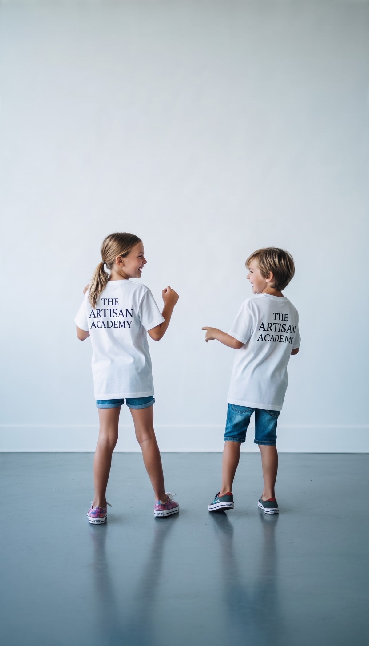 A young girl and a boy viewed from behind, captured in a candid, playful moment indoors. Both wear matching white crewneck t-shirts featuring the stacked dark text "THE ARTISAN ACADEMY" on the upper back. The girl on the left, with her hair in a ponytail, wearing denim shorts and patterned pink sneakers, gestures towards the boy on the right, who wears longer denim shorts and dark sneakers as they laugh together.