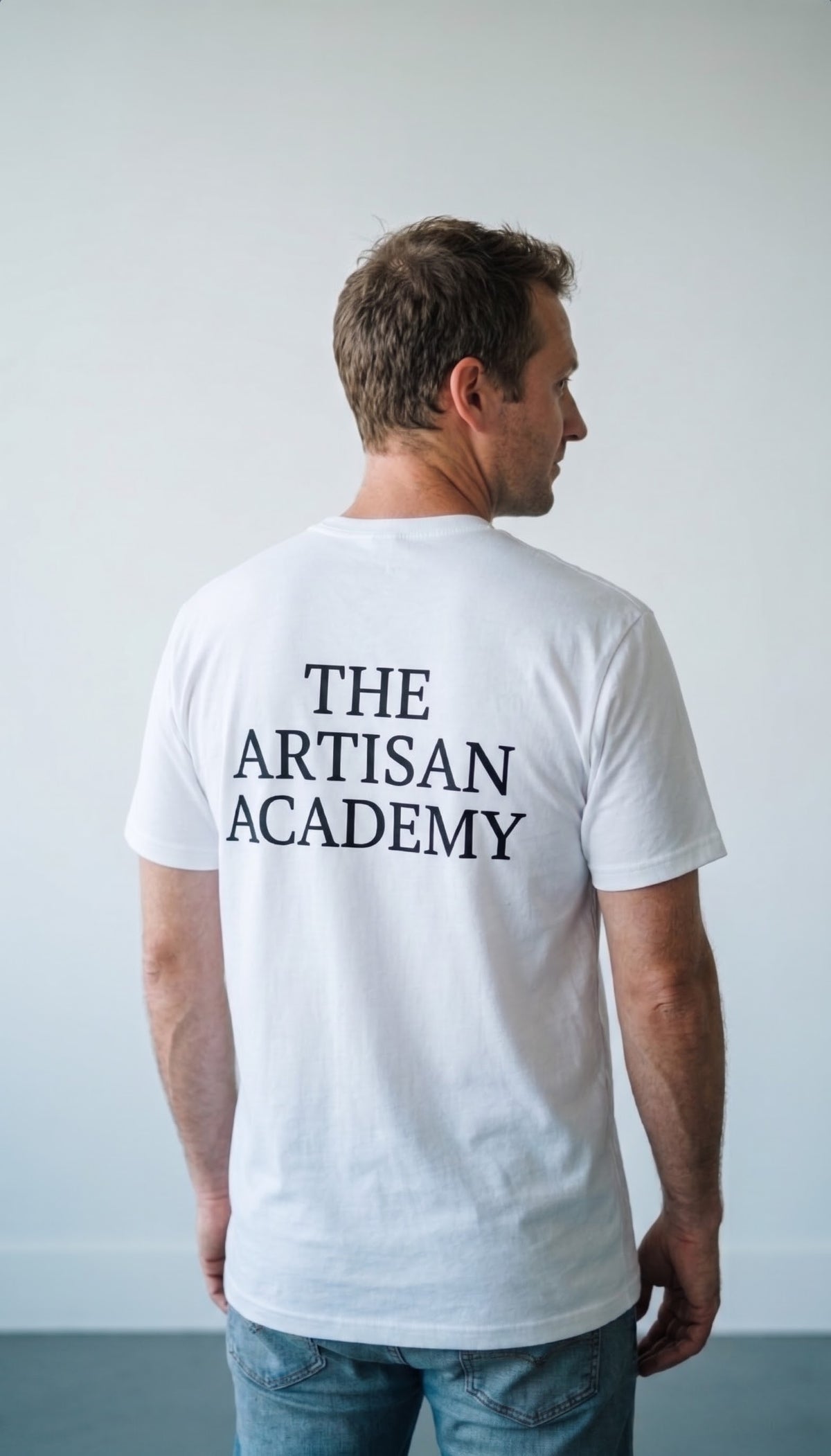 A candid, close-up shot of a man viewed from behind and slightly to the side, standing in a minimalist indoor studio. He wears a white crewneck t-shirt featuring the large, bold, stacked black text "THE ARTISAN ACADEMY" across the upper back, paired with blue jeans. He has short brown hair and is looking slightly to his right, captured from the waist up.