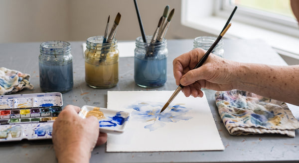 A close-up of a person's hands painting on a white sheet of paper with blue and yellow watercolour, surrounded by paint jars, brushes, and a watercolour palette on a grey table.