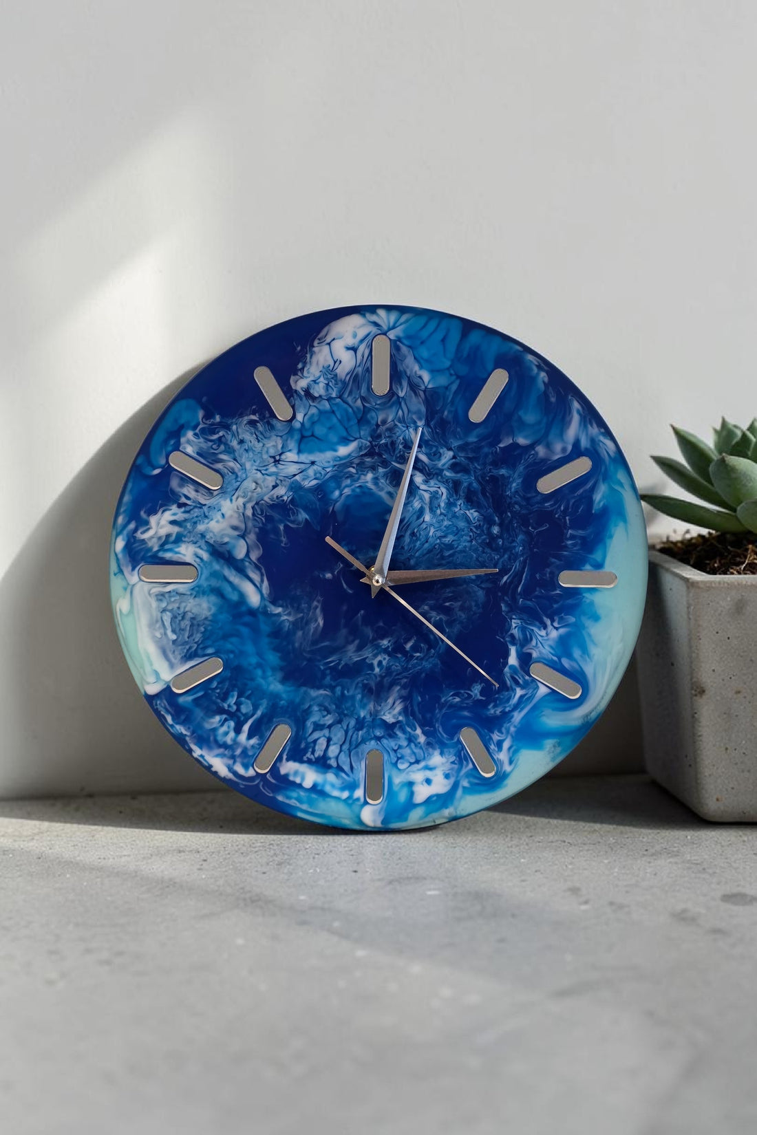 Front face close-up of a circular handmade wall clock featuring vibrant blue and white fluid resin art, styled standing upright on a grey concrete shelf against a minimalist white wall. The clock displays silver hands over deep azure, teal, and seafoam cells, with vertical cutout slots serving as hour indicators.