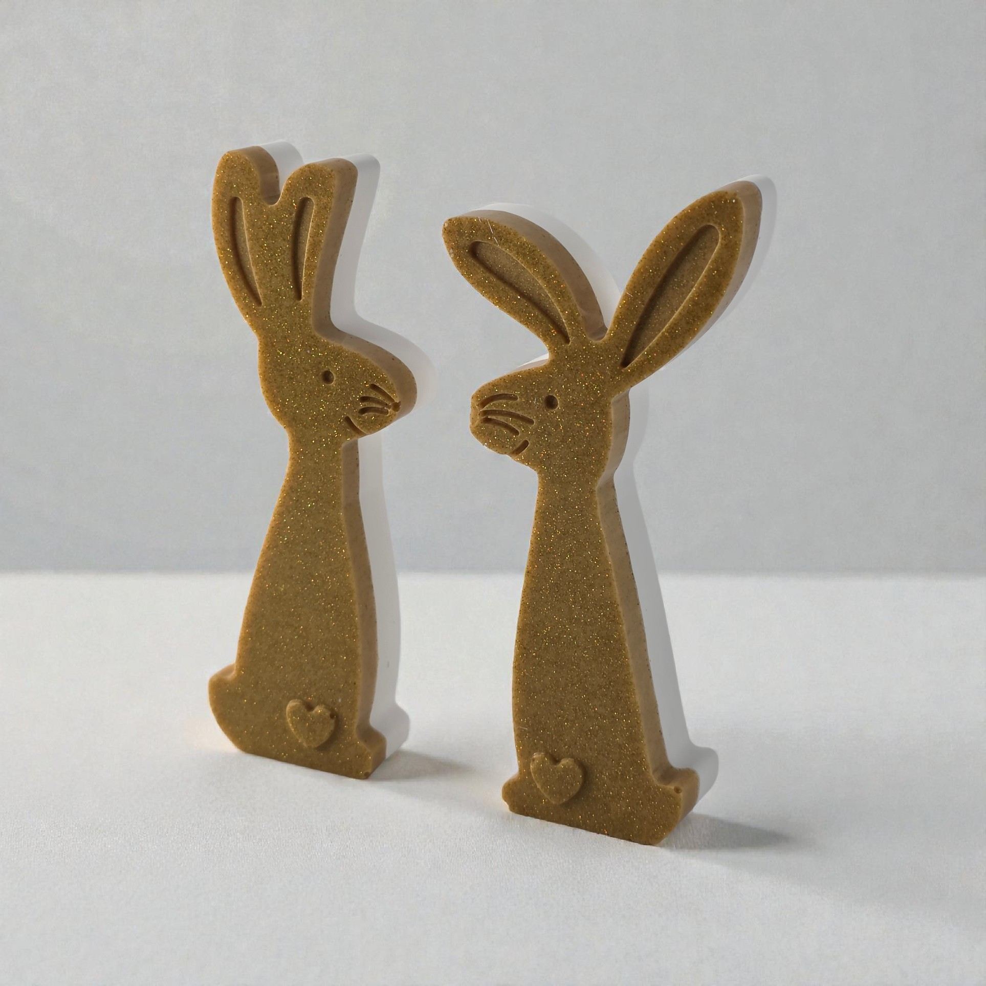 Two slim, golden glitter bunny figurines with small heart details, standing on a white surface against a light background.