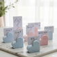 Hand-poured heart-shaped matte stone card holders in pastel pink and blue displaying wedding place cards and retail price tags on a marble surface.