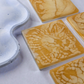 A top-down view of four square gold resin coasters with organic white and honey-toned swirling patterns on a textured grey background.