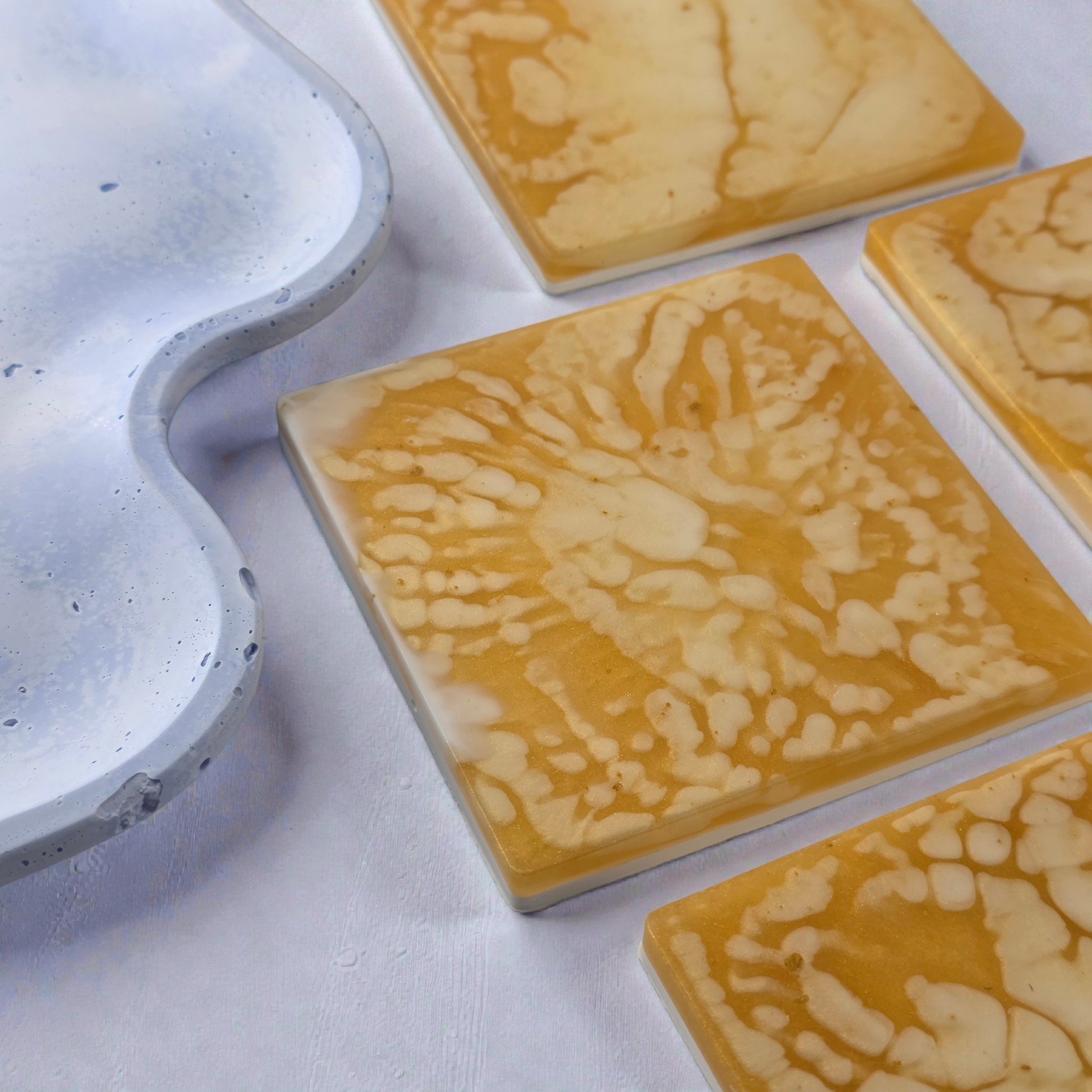 A top-down view of four square gold resin coasters with organic white and honey-toned swirling patterns on a textured grey background.