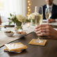 A lifestyle shot of the golden coasters in use at a formal dinner table, featuring champagne glasses and white floral arrangements.
