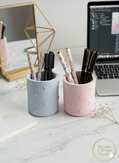 A pair of matte stone pen cups in pastel blue and pink with embossed heart patterns on a white marble desk next to a laptop