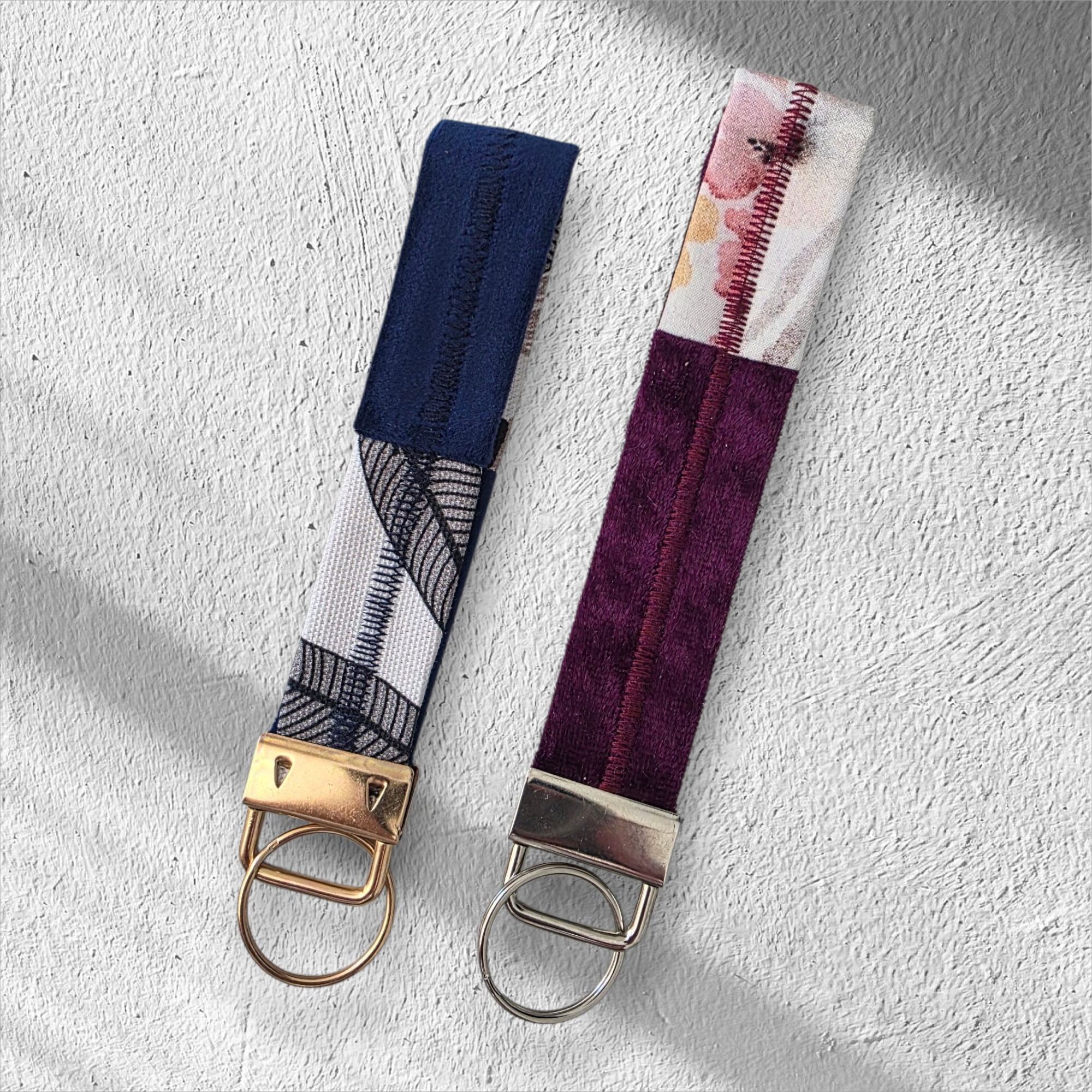 Two fabric keychains side-by-side: one navy and white with gold hardware, and one purple and floral with silver hardware.